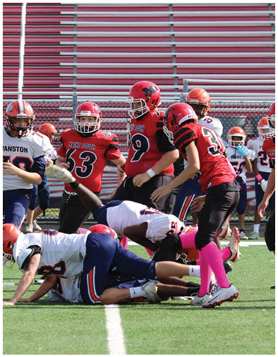 People wearing football uniforms are engaged in a play on a green field with red bleachers in the background. Description generated by AI