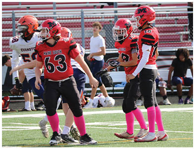 Several people wearing red and black football uniforms with pink socks are standing on a sports field with bleachers and other people in the background. Description generated by AI