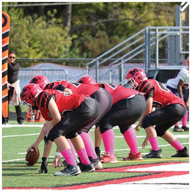A group of children in red football uniforms and pink socks are lined up on a football field preparing for a play while a child in a white uniform stands in the background. Description generated by AI