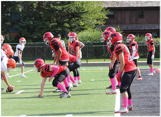 Two youth football teams in red and white uniforms face each other on a green field with one team preparing to snap the ball. Description generated by AI