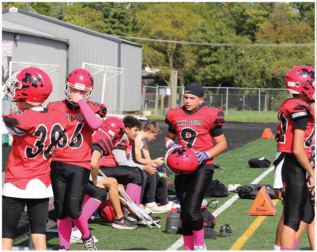 People wearing red and black football uniforms are gathered on a sports field with some sitting on a bench and others standing or walking. Description generated by AI