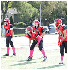 Four people in red and black football uniforms with pink socks and gloves stand on a grassy field preparing for a play. Description generated by AI
