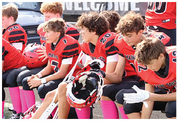 A group of children wearing red and black football uniforms with pink socks are sitting on a bench, some holding helmets and crutches. Description generated by AI