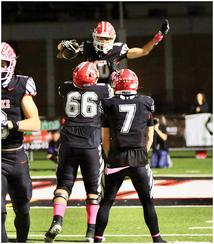 Four people in black football uniforms with red helmets are on a field, with one person jumping and others standing or walking. Description generated by AI