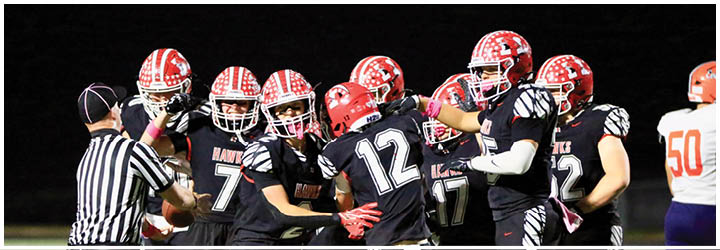 A group of people wearing black and red football uniforms are celebrating on a field while a referee signals nearby and a person in an orange and white uniform walks away. Description generated by AI