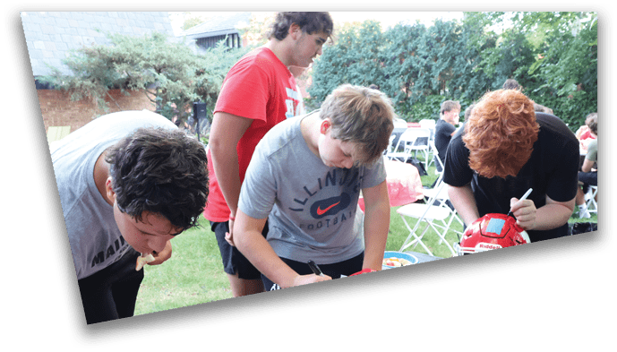 Three people are outdoors at a table with a red cloth, with one person writing on a paper, another leaning over the table, and a third holding a red football helmet. Description generated by AI