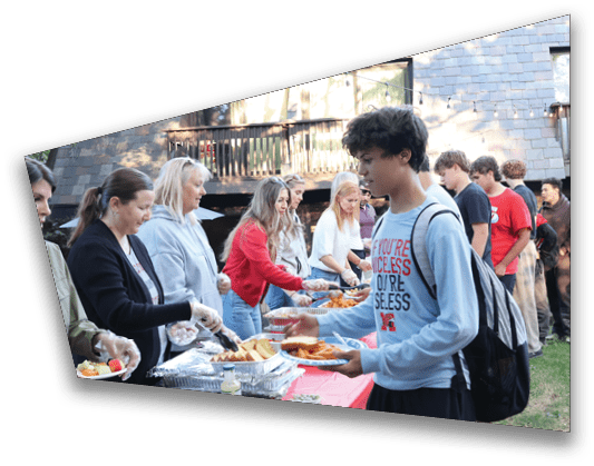 People are gathered outdoors in a backyard setting, standing in line to receive food from a buffet table with various dishes. Description generated by AI
