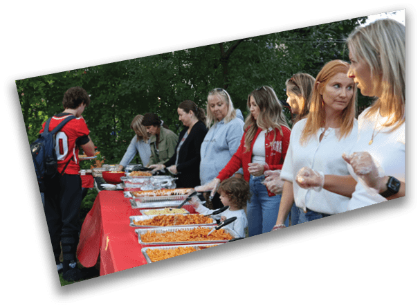 People stand in line outdoors at a buffet table with trays of food while one person in a sports jersey with a backpack serves themselves. Description generated by AI