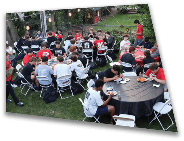 A large group of people wearing casual clothing are seated outdoors on folding chairs around tables, eating and socializing under string lights in a grassy backyard setting. Description generated by AI