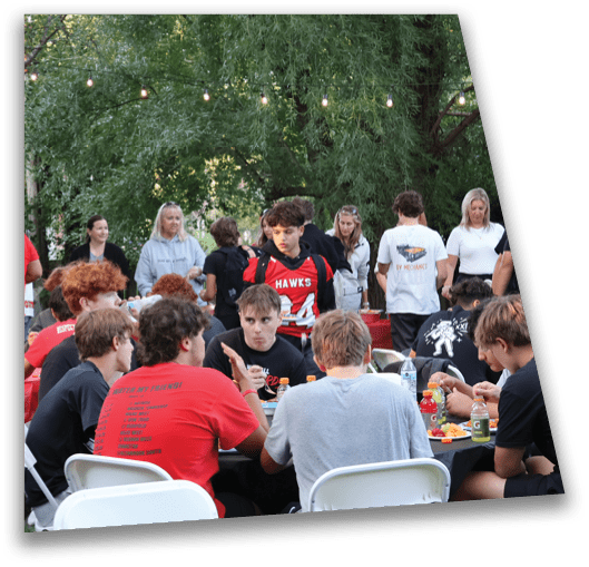 A group of people wearing casual clothing are gathered outdoors around tables with red tablecloths, eating and socializing under string lights and trees. Description generated by AI