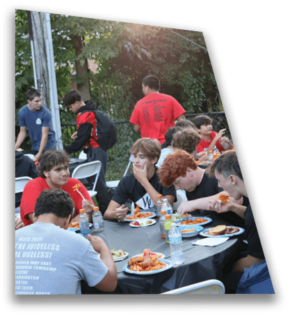 A group of people wearing casual clothing are sitting and standing around tables outdoors, eating food and socializing in a leafy setting. Description generated by AI