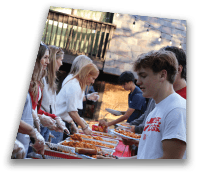 People wearing casual clothes serve food from trays to others holding plates in an outdoor setting with a wooden fence and building in the background. Description generated by AI