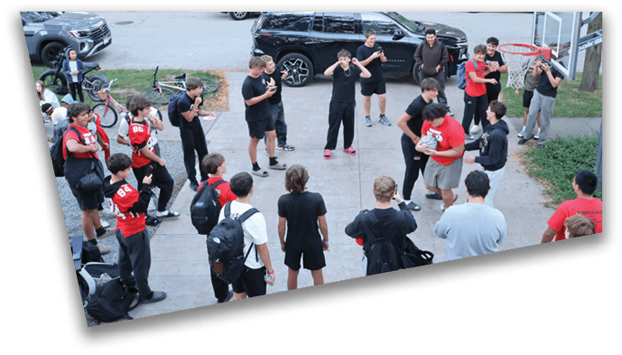 A group of people wearing casual and sports clothing are gathered on a driveway near a basketball hoop, with some standing in a circle and others watching. Description generated by AI