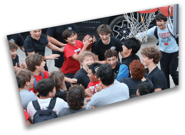 A group of excited people wearing casual clothes are gathered closely around a basketball hoop, with one person holding a basketball and others reaching toward it. Description generated by AI