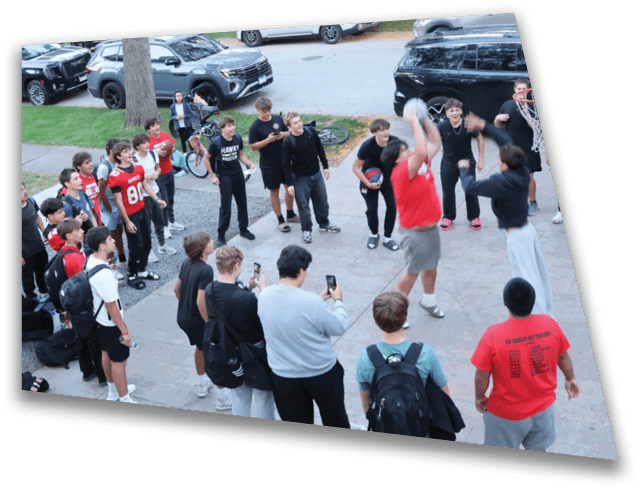A group of people gathered outdoors on a paved area with some watching and others actively playing basketball while several vehicles are parked nearby on the street. Description generated by AI