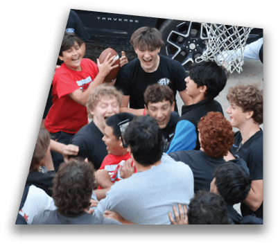 A group of people wearing casual clothes are gathered closely outdoors around a basketball hoop, smiling and holding a basketball. Description generated by AI