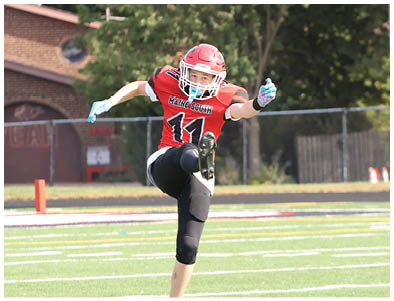 A person wearing a red football uniform and helmet is kicking a football on a grassy field with trees and buildings in the background. Description generated by AI