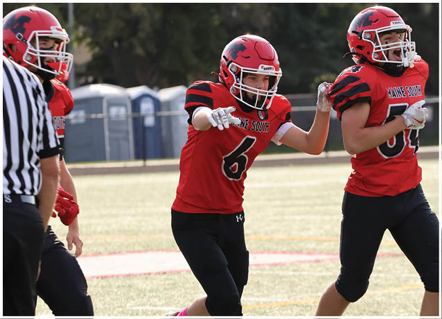 Two people in red football uniforms and helmets are on a field with one pointing forward and the other running beside them. Description generated by AI