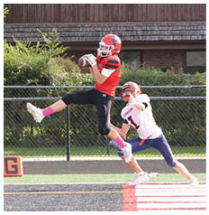 Two people in football gear are actively competing near the end zone on a field with a house and portable toilets in the background. Description generated by AI