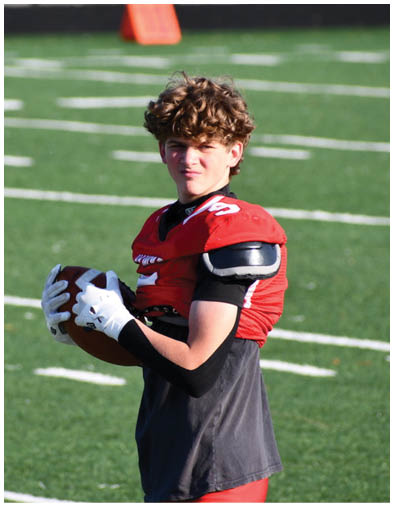 A person with curly hair wearing a red football jersey and gloves is holding a football on a grassy field. Description generated by AI