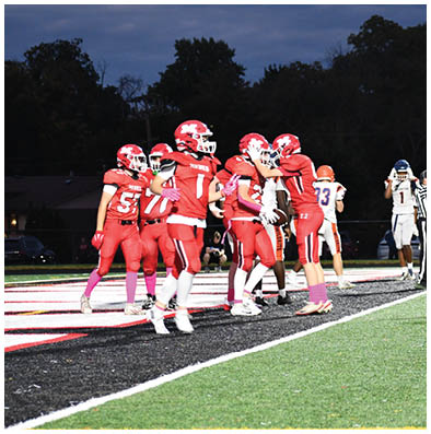 A group of people in red football uniforms are celebrating on a field near the end zone while a referee signals and opposing players stand nearby. Description generated by AI