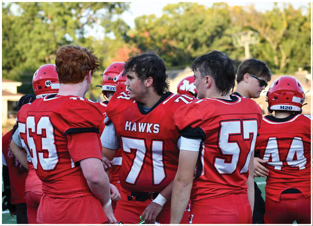 A group of people wearing red football uniforms with numbers and the word Hawks are gathered outdoors, some facing each other and others looking away. Description generated by AI