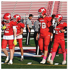 Four people in red football uniforms with helmets stand on a football field near referees in striped shirts and empty bleachers. Description generated by AI