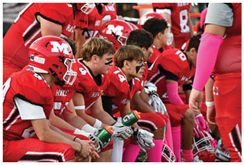 People in red football uniforms with black face paint sit on a bench holding water bottles and wearing pink accessories. Description generated by AI