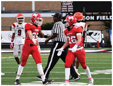 Two people in red football uniforms are on a field with a referee walking between them and two other players in the background. Description generated by AI