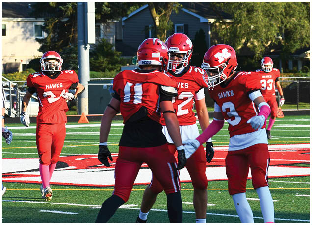 Four people in red football uniforms are on a field with one person running in the background and three standing together in the foreground. Description generated by AI
