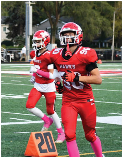 Two people in red football uniforms with pink socks and gloves are running on a green football field near an orange marker with the number 20. Description generated by AI