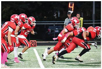 Several athletes in red football uniforms are lined up and poised to start a play on a football field with a down marker showing the number four in the background. Description generated by AI