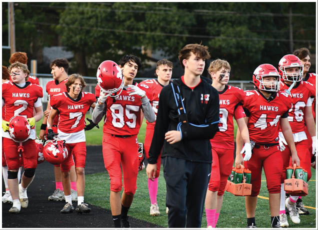 A group of people in red football uniforms and helmets are walking together on a field, with one person in a black jacket and arm sling walking in front. Description generated by AI