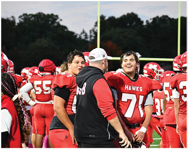 People in red football uniforms and a coach in a black and red jacket are gathered on a field, with one player smiling and interacting with the coach. Description generated by AI