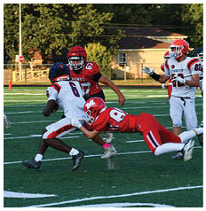 Four people wearing red helmets and white football jerseys with red stripes stand in a row on a sports field during daylight. Description generated by AI