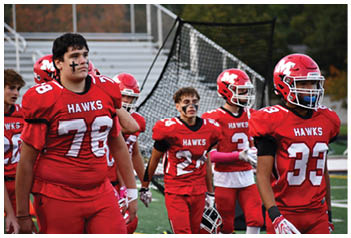 Several adults wearing red football uniforms with “HAWKS" on the front and black face paint are standing on a sports field near a goal net. Description generated by AI