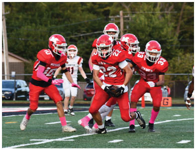 Several people in red football uniforms are actively playing on a field with a few people in white uniforms in the background. Description generated by AI