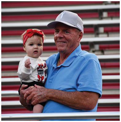 A senior wearing a blue polo shirt and gray cap is holding a child dressed in a white shirt with a red bow headband while standing in front of red and white bleachers. Description generated by AI