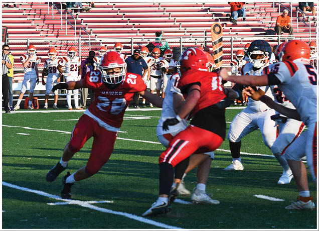 Players in red and white football uniforms are actively engaged in a play on a green field with empty bleachers and a few spectators in the background. Description generated by AI
