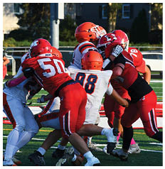 Several people wearing football uniforms in red and white are engaged in a close physical play on a green field during a game. Description generated by AI