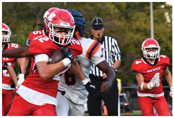 A group of people in football uniforms are actively playing on a field with one person in red running with the ball while others in red and white uniforms chase or block. Description generated by AI