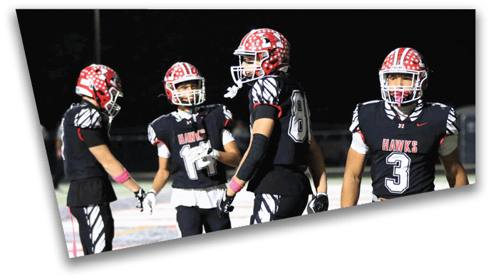 Several people in black football uniforms with “HAWKS" on the front are standing on a football field near the end zone at night. Description generated by AI