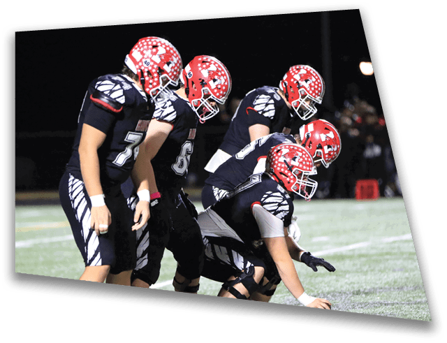 Four people in football uniforms and red helmets are lined up on a field with one person holding a football ready to snap it. Description generated by AI