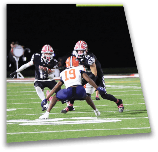 Two people in black sports uniforms with red and white helmets are running toward a person in a white and orange sports uniform on a football field near a goalpost at night. Description generated by AI