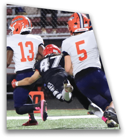Three football players in helmets and uniforms are actively engaged in a play on the field, with one player in black diving toward the ground while two players in white and orange move nearby. Description generated by AI