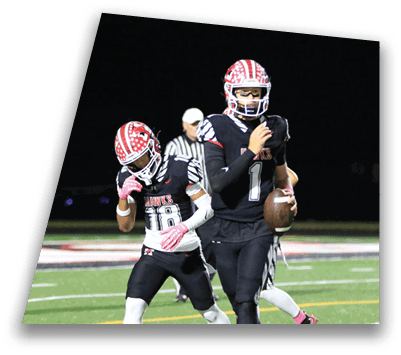 Two people in football uniforms and helmets are on a field at night, with one holding a football and the other adjusting their helmet. Description generated by AI