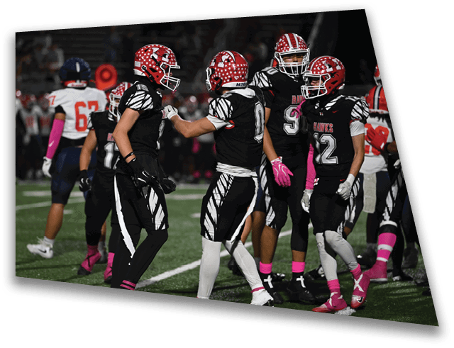 Two people in black and white football uniforms with red helmets are interacting on a football field while other players stand in the background. Description generated by AI