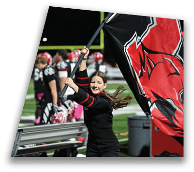 A person wearing a black outfit is smiling and waving a large red and black flag on a sports field with players in football uniforms standing in the background. Description generated by AI