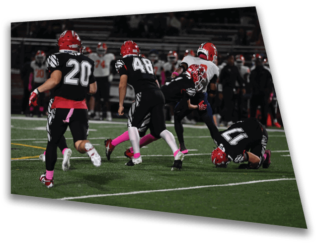 Several people wearing football uniforms are on a field with one person on the ground holding a football while others are running nearby. Description generated by AI