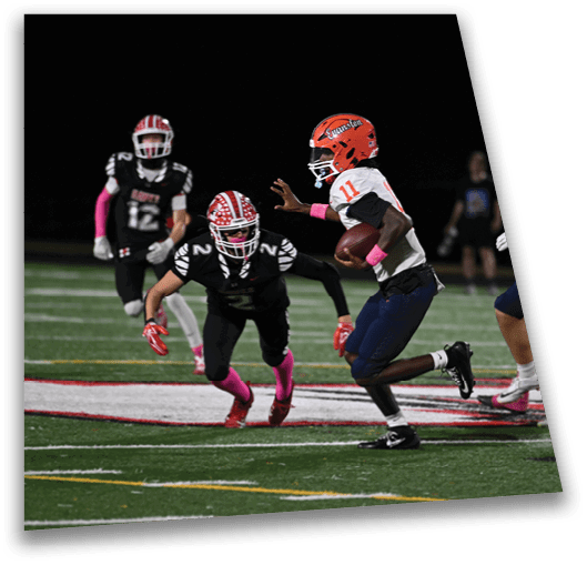 Adults in football uniforms are playing a nighttime game on a field with one player in an orange helmet running with the ball while others in dark and white uniforms move around him. Description generated by AI